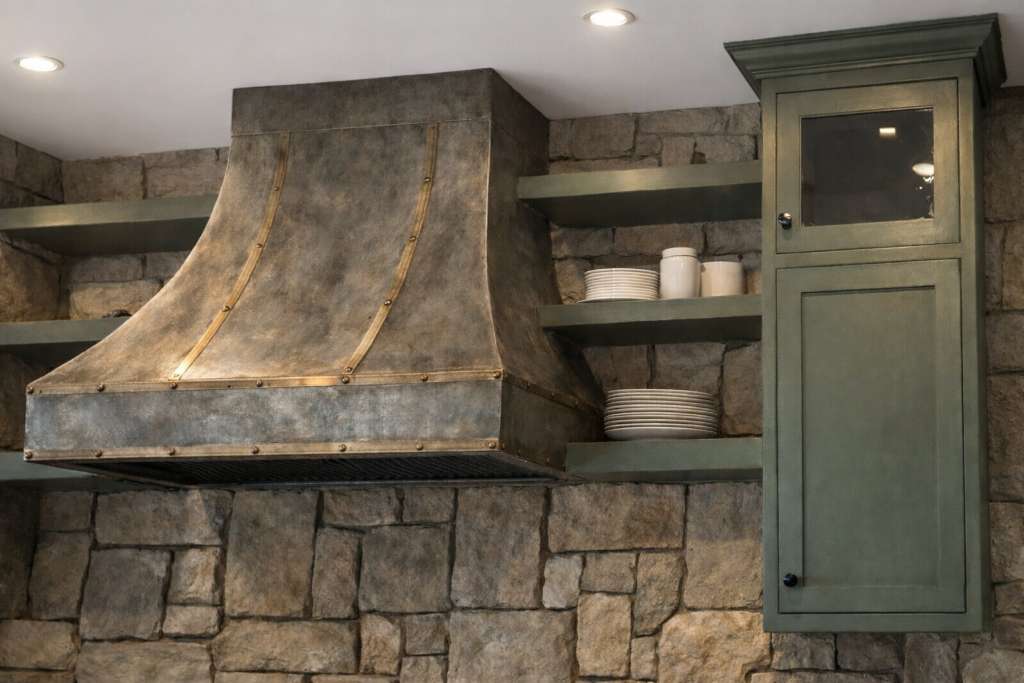 Rustic kitchen detail with a patinated metal range hood, brass straps, sage-green cabinetry, open shelves with white dishware, and a rough-cut stone backsplash beneath a solid white ceiling.