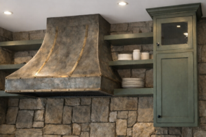 Rustic kitchen detail with a patinated metal range hood, brass straps, sage-green cabinetry, open shelves with white dishware, and a rough-cut stone backsplash beneath a solid white ceiling.